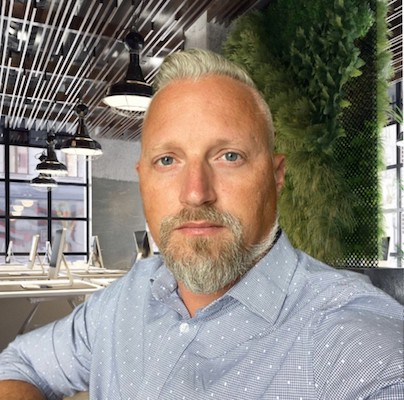 White man with a blue button up with plants in the backgound