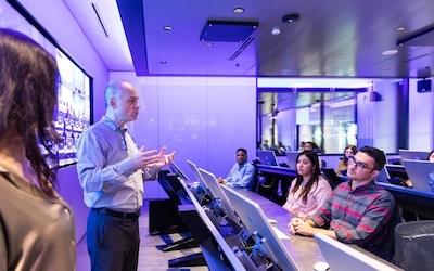 A professor teaching at the front of a class in a computer room