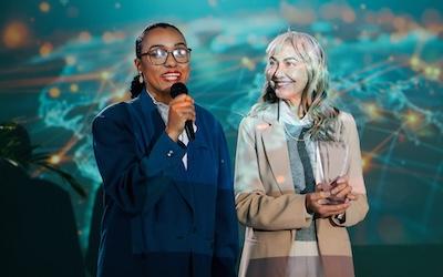 two business woman; one black and the other white talking into microphones while a projector shines on them