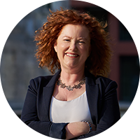 headshot of a professional business woman with auburn hair smiling, she is wearing a black suit, white blouse and a necklace
